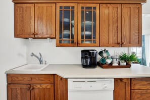 A kitchen with wooden cabinets, a white dishwasher, a coffee maker, and a plant on the counter. - Heated Pool Access | 5*Clean | Flex Cancelation (Tybee Island)