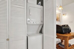 A compact, white laundry room with a built-in washer and dryer, a wooden table with a radio, and a framed picture on the wall. - Heated Pool Access | 5*Clean | Flex Cancelation (Tybee Island)