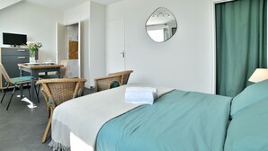 Habitación de hotel con cama, escritorio con computadora, dos sillas, una silla de mimbre, un espejo y una cortina verde.