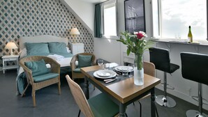 Una habitación con una mesa de comedor preparada para dos personas, un sofá con cojines verdes, una silla de mimbre y una planta en maceta con flores.