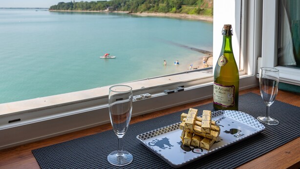 Vista desde una ventana a una playa con una botella de vino, una copa y un plato de tapones de corcho sobre una alfombra.