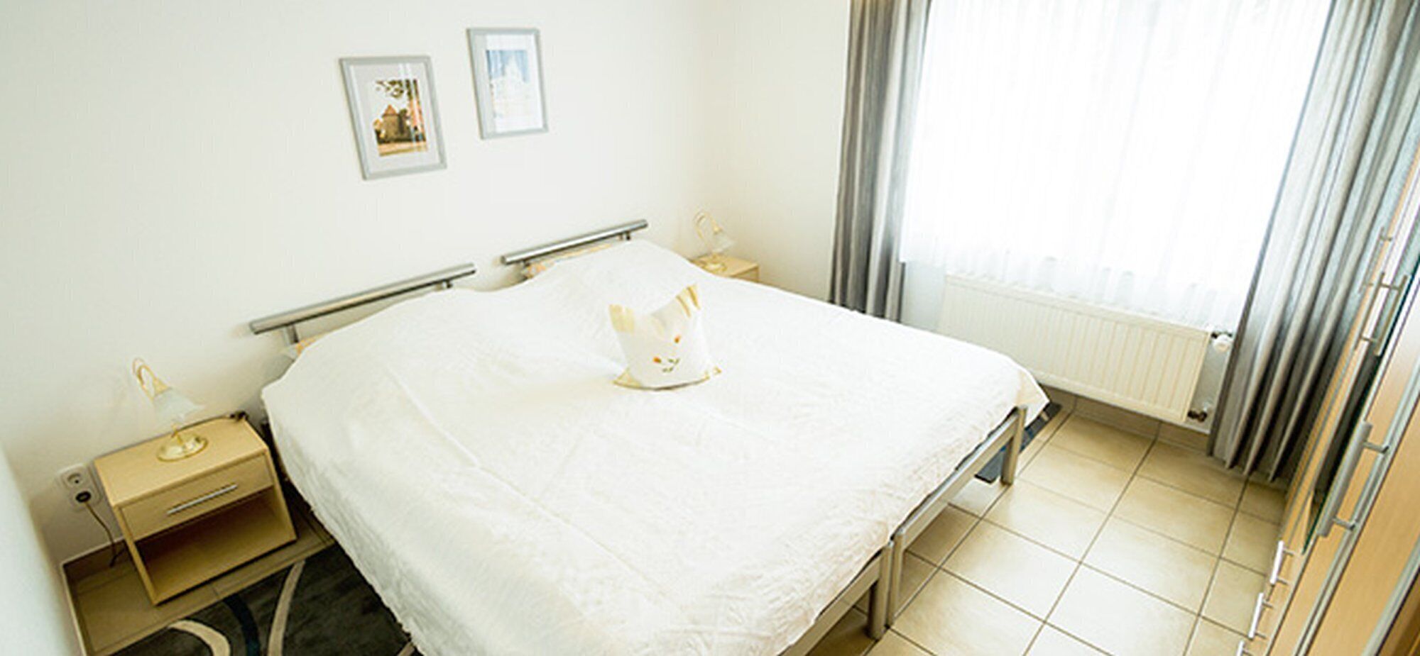 Et hotelværelse med seng, sengeborde, en radiator og et vindue med gardiner.