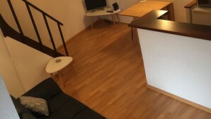 Een knus woongedeelte met een houten vloer, een zwarte bank met kussens, een klein wit tafeltje, een televisie en een magnetron.