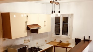 Een kleine keuken met een inbouwmagnetron, fornuis en spoelbak. Er is een raam met zonwering en een hanglamp boven het aanrecht.