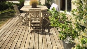 Una terraza de madera con una mesa y sillas, rodeada de plantas en macetas y un arbusto blanco con flores.