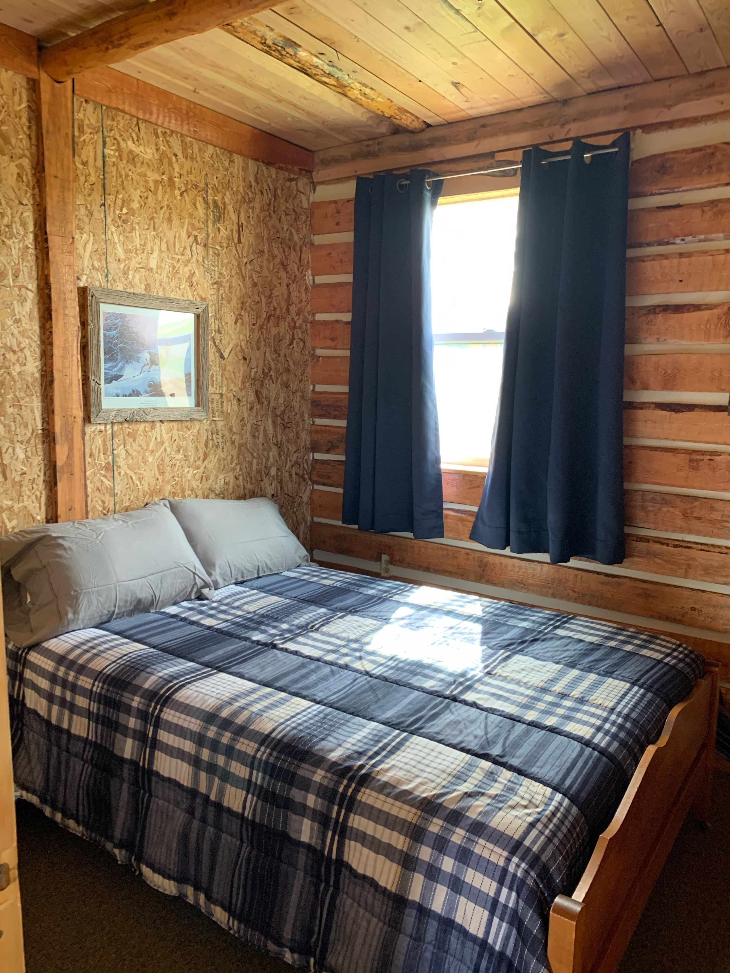 Une chambre dans une cabane en bois, avec une couverture à carreaux sur le lit, un tableau encadré au mur et une fenêtre avec des rideaux.