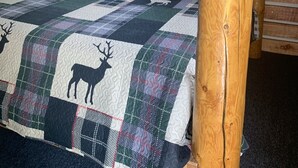 Habitación de cabaña de troncos con una cama que incluye un colcha con motivos de ciervo, almohadones a cuadros y un cuadro enmarcado con animales salvajes en la pared.