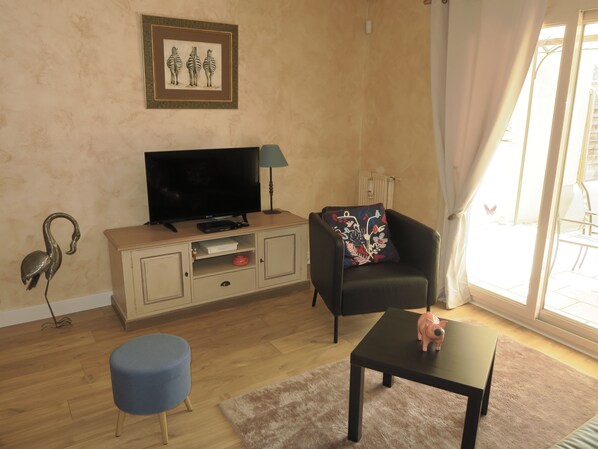 A living room with a flat-screen TV on a wooden cabinet, a black armchair with a floral pillow, a small blue ottoman, and a piggy bank on a table.