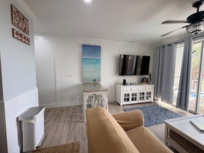 Een woonkamer met een beige bank, een kleine tafel, een televisie en een raam met gordijnen.