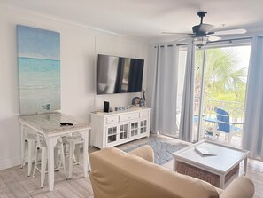 Una sala de estar con televisor, muebles blancos, un balcón con sillas y vistas al exterior.