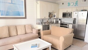 A living room with a beige sofa, a white coffee table, and a kitchen with white cabinets and stainless steel appliances. - Coastal Condo with Amazing View & Beach Access (Santa Rosa Beach)