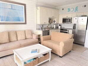 Una sala de estar con un sofá color beige, una mesa de centro blanca y una cocina con gabinetes blancos y electrodomésticos de acero inoxidable.