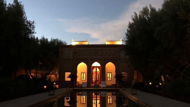 Una piscina con el reflejo de un edificio con ventanas de arco y una puerta central, rodeado de árboles y un camino empedrado.