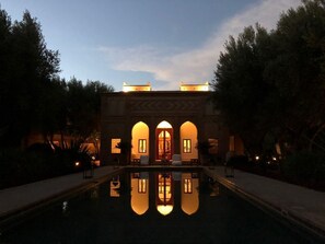 Una piscina amb el reflex d'un edifici amb finestres allargades i una porta central, envoltat d'arbres i un passeig pavimentat.