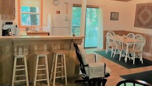 A kitchen with a bar area, white bar stools, a dining table with white chairs, and a view of the outdoors. - Riverside Retreat  (Elkins)