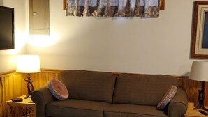 A living room with a brown sofa, a patterned rug, a wooden floor, a window with curtains, a wall-mounted TV, and a framed picture. - Riverside Retreat  (Elkins)