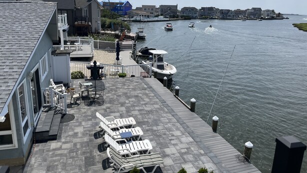 A waterfront patio with lounge chairs and a dining area, overlooking a calm river with boats. - BEST BAYFRONT VALUE IN CHADWICK BEACH NEWLY RENOVATED SLEEPS 7 RATED 10/10 (Lavallette)