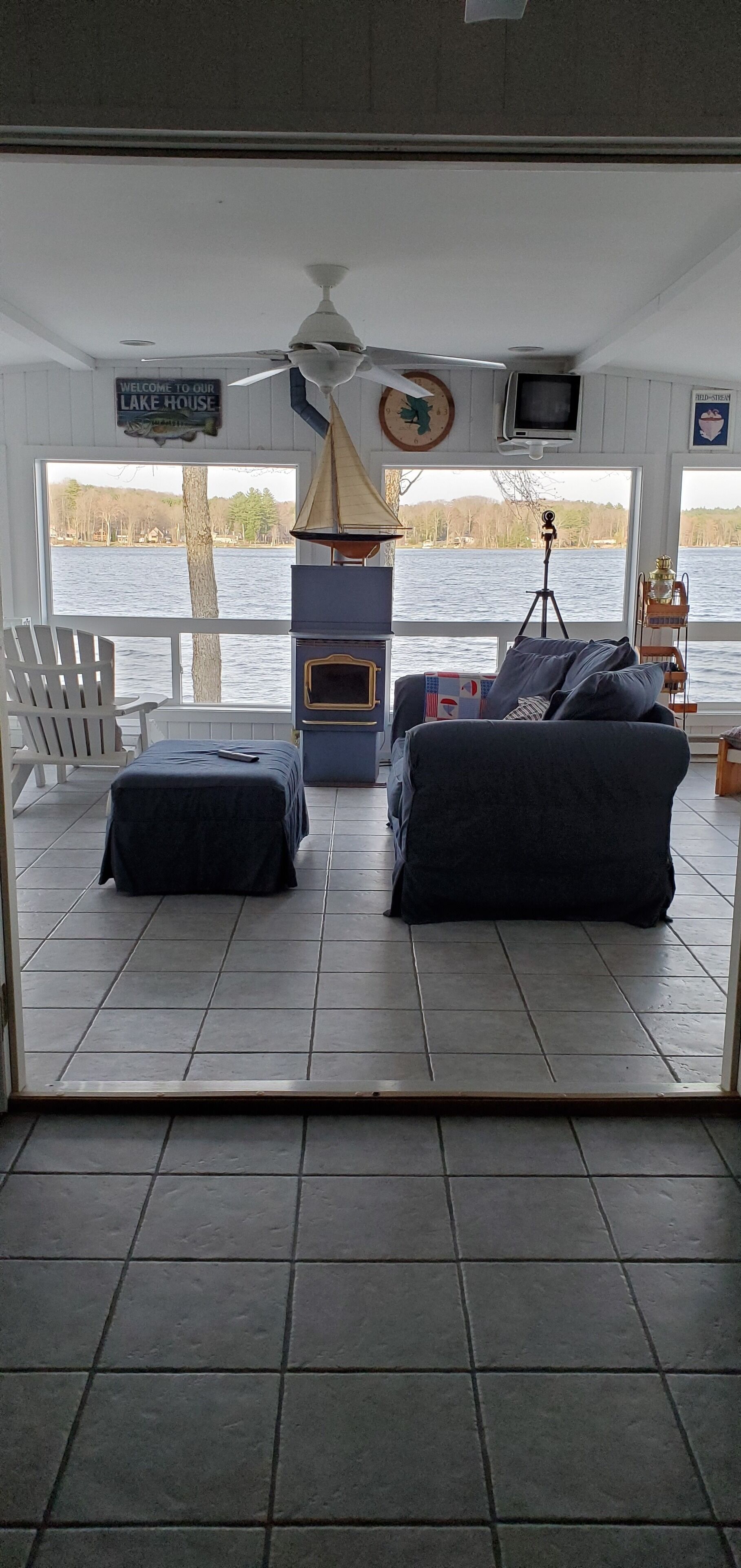 A room with a view of a lake, a ceiling fan, a fireplace, and a sign that reads "Welcome to our Lake House."