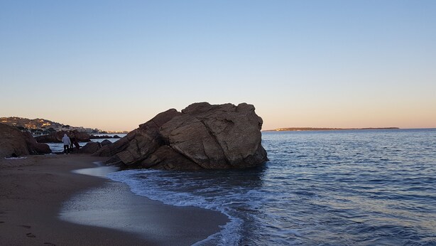 Ein felsiger Strand mit einem großen Felsen, ruhigem Meer und einer fernen Küste mit Gebäuden.