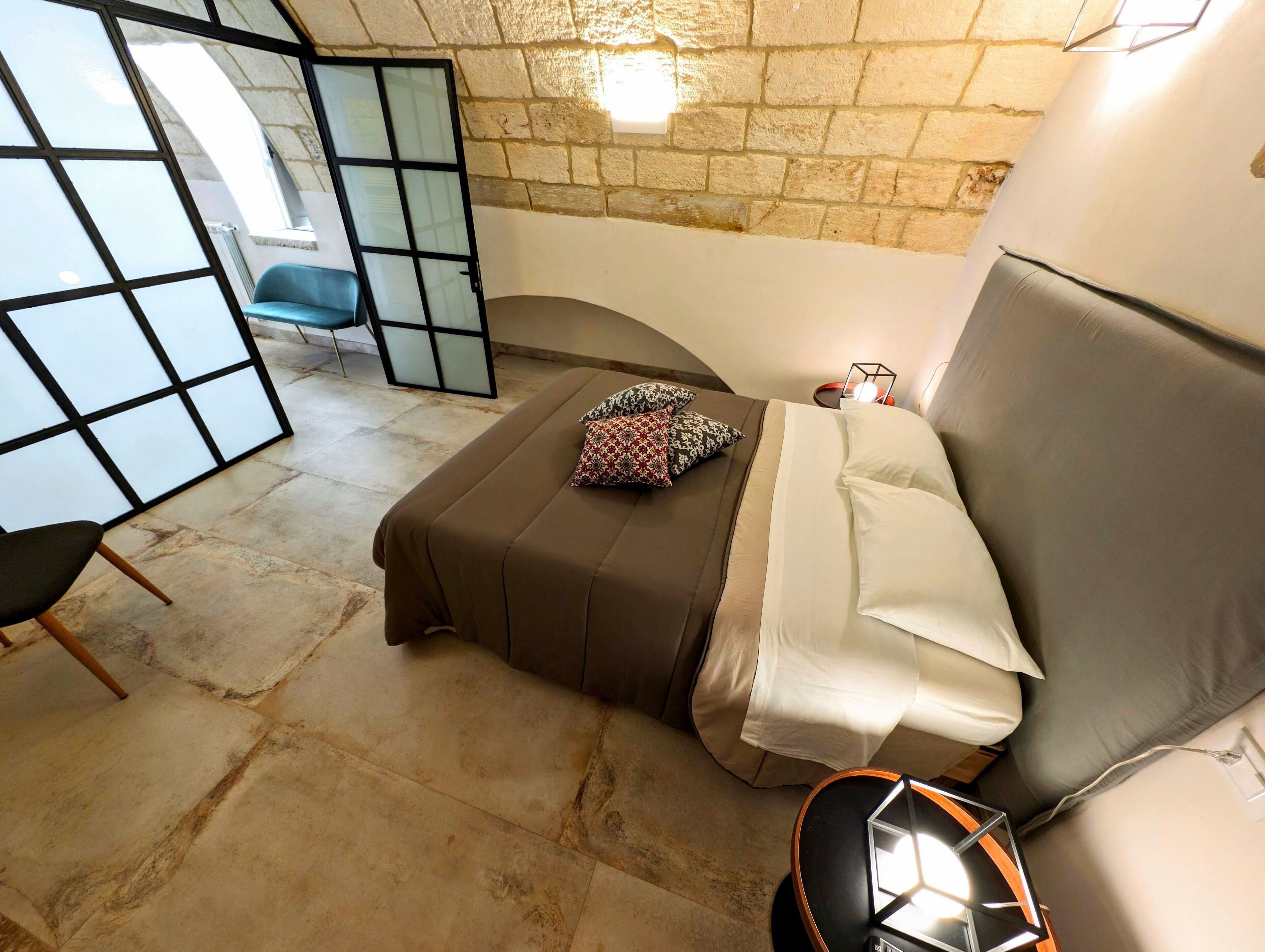 A cozy bedroom with a bed, a chair, a small table, and a stone wall.