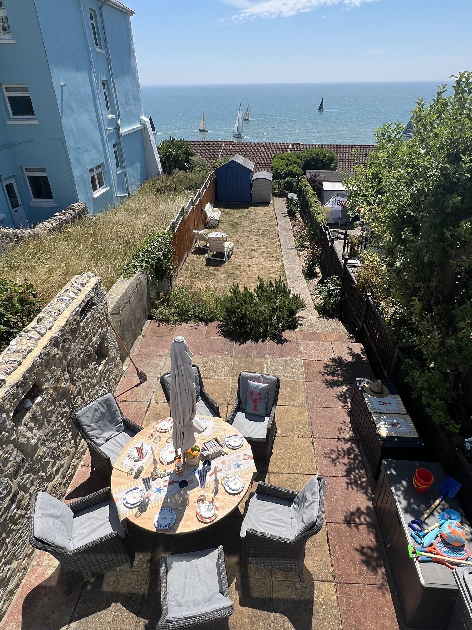 Een terras met een ronde eettafel, omringd door rieten stoelen, met uitzicht op zee en zeilboten.