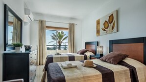 Habitación de hotel con dos camas, un aparador, un espejo, una planta y un balcón con vistas.