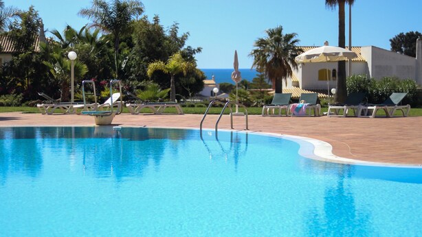 Una piscina cristalina con sillas de descanso y sombrillas, rodeada de palmeras y un edificio con tejado de baldosas.