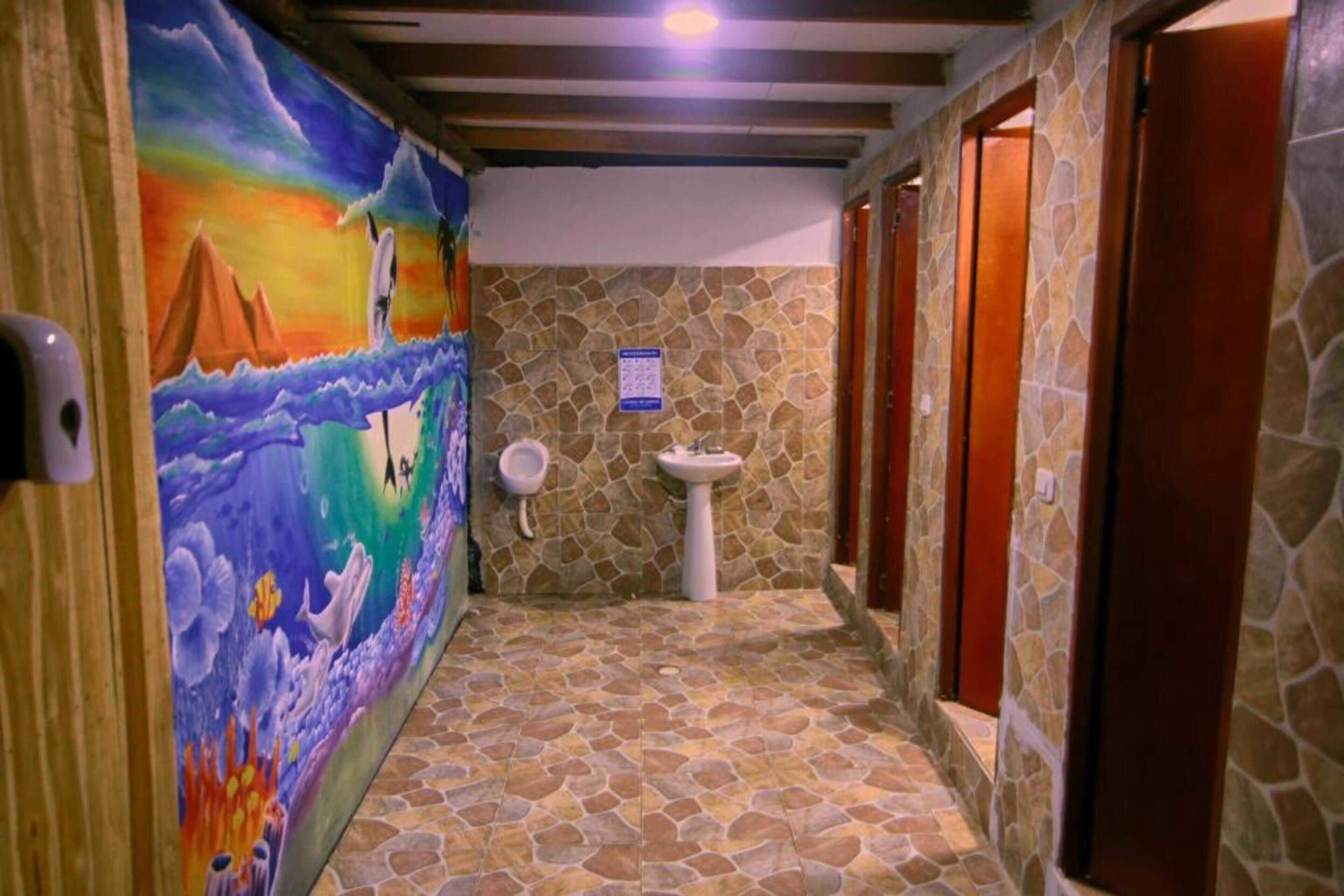 Un pasillo con suelo de piedra, un mural en la pared izquierda, un lavabo y puertas de madera.