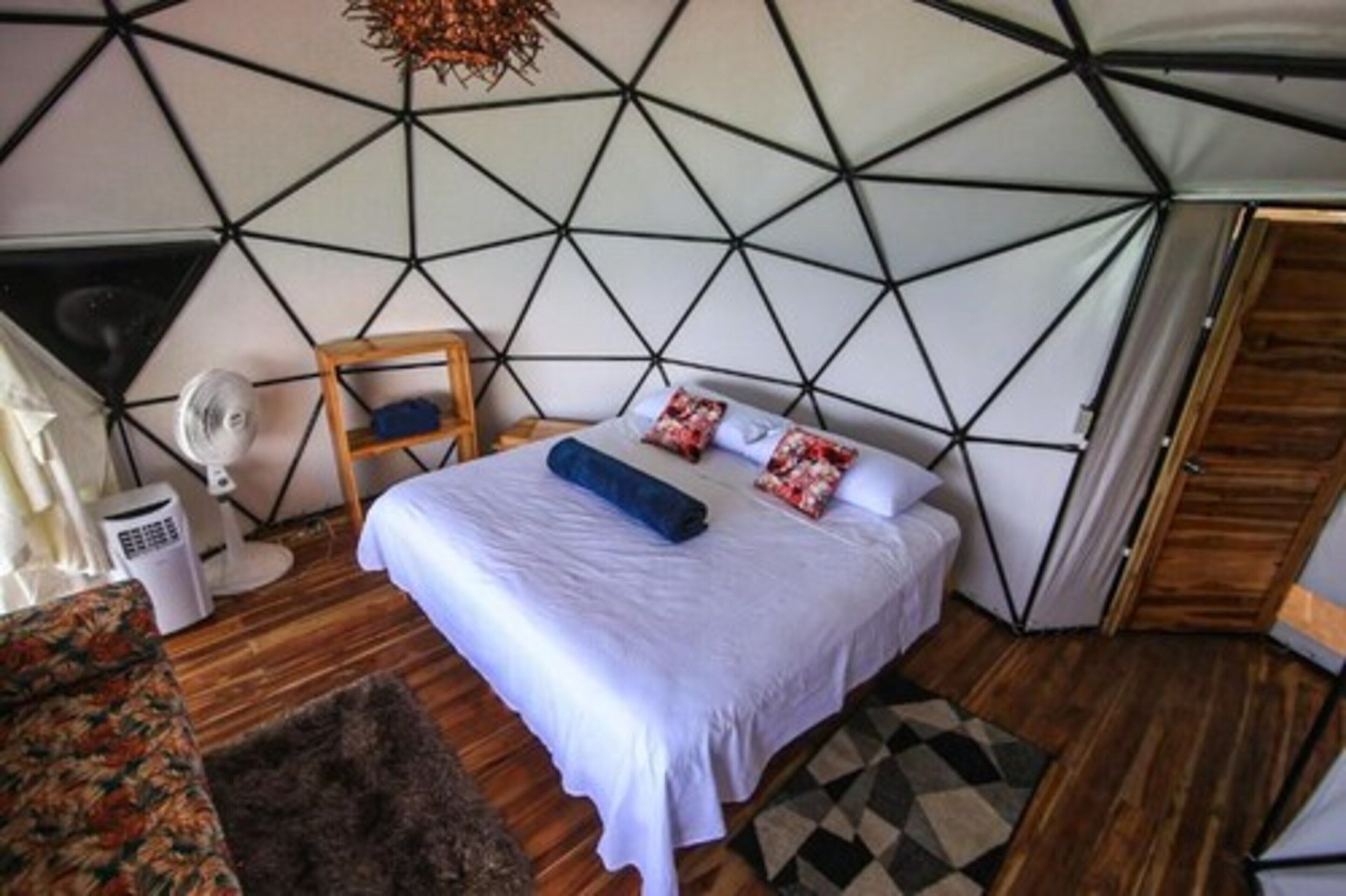 Un dormitorio con techo estampado con motivos geométricos, una cama con ropa blanca, una silla de madera, un ventilador y una alfombra pequeña.