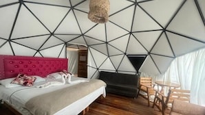 A modern bedroom with a geometric ceiling design, a tufted pink headboard, a bed with white linens, a dark sofa, a small round table, and a patterned rug. - Cabana Glamping Geronimo  (Cartagena)