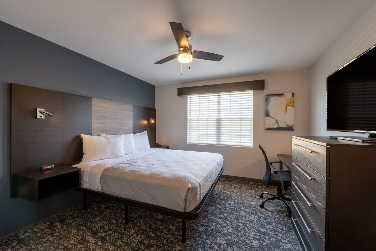 Habitación de hotel moderna con cama, escritorio, silla, cómoda, televisión, ventilador de techo y ventana con persianas.
