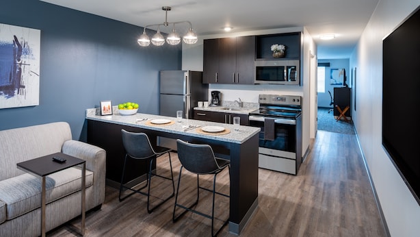 A modern kitchen with a central island, dark cabinetry, stainless steel appliances, and a seating area with a sofa and chairs. - Stay APT Suites Alexandria-Fort Belvoir (Alexandria)