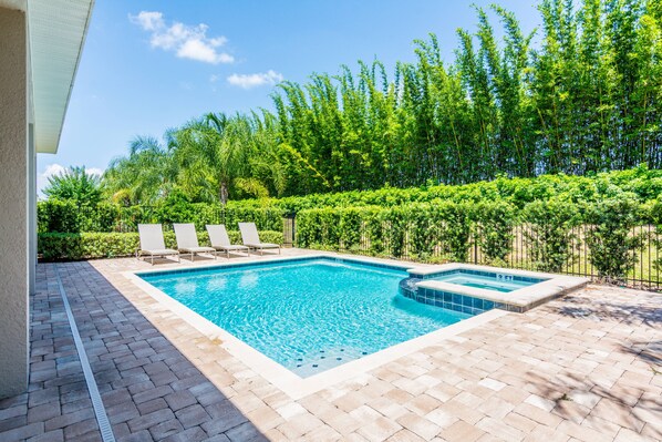 Una piscina rectangular con un jacuzzi, rodeada por un patio de ladrillo y sillas de descanso blancas.