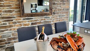 En stenmur med ett kök synligt genom ett fönster, ett bord med en hink champagne och en skål med skaldjur, och en matplats med stolar.