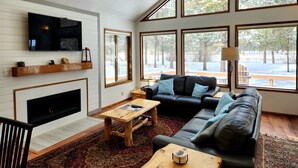 Una sala de estar moderna con chimenea, ventanas amplias, un sofá de cuero negro y una mesa de centro de madera.