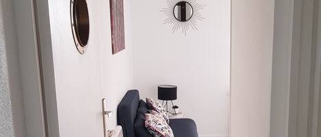 A room with a dark blue sofa, a round mirror, a small table with a lamp, and a wall-mounted sunburst decoration.
