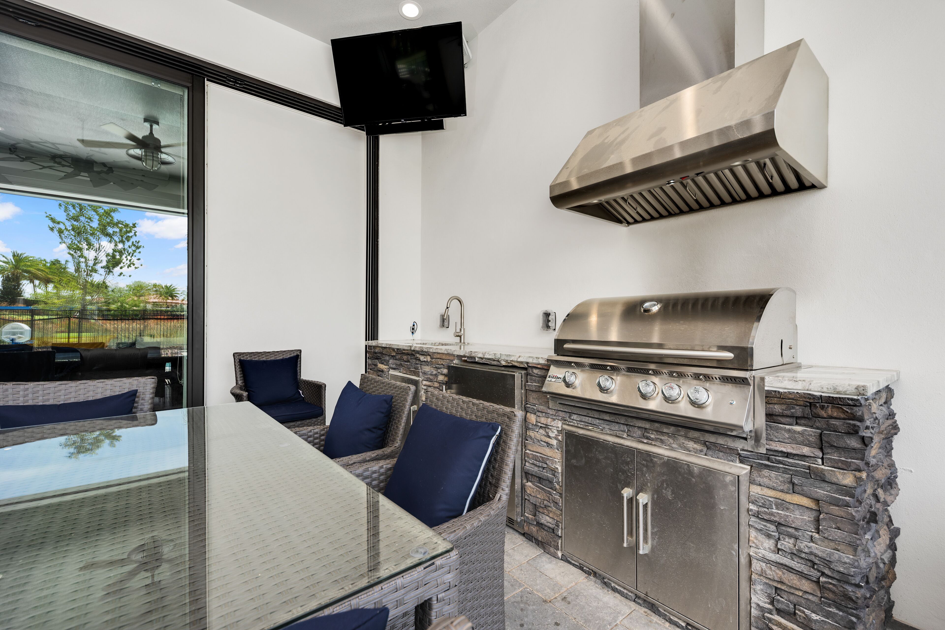 An outdoor kitchen with a stone backsplash, a stainless steel grill, and a glass dining table with blue cushions.