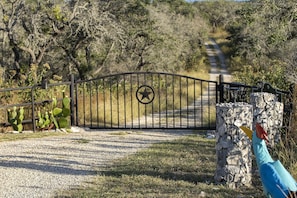 Enter through the gated entrance, flanked by a stone pillar and a cactus, onto a gravel path leading to a dirt road.