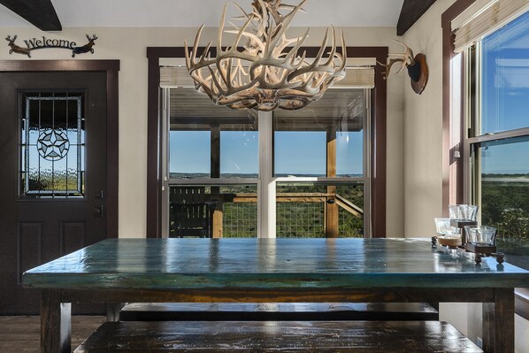 Experience a welcoming atmosphere in this cozy space, featuring a rustic table, unique chandelier, and stunning views through large windows.