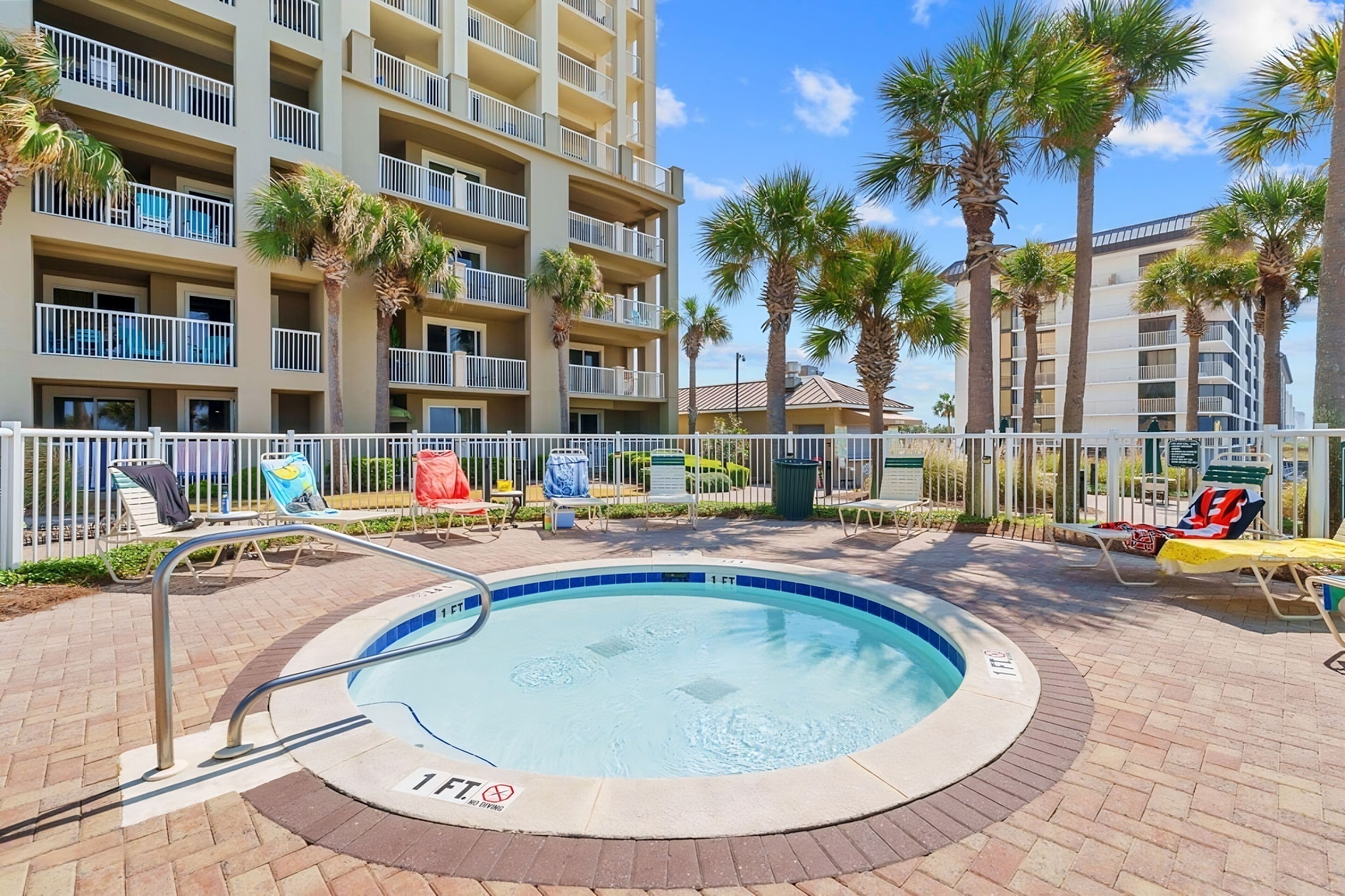 A pool area with lounge chairs, palm trees, and a multi-story building with balconies.
