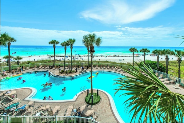 A pool with a curved edge, surrounded by lounge chairs and palm trees, with a beach and ocean in the background.