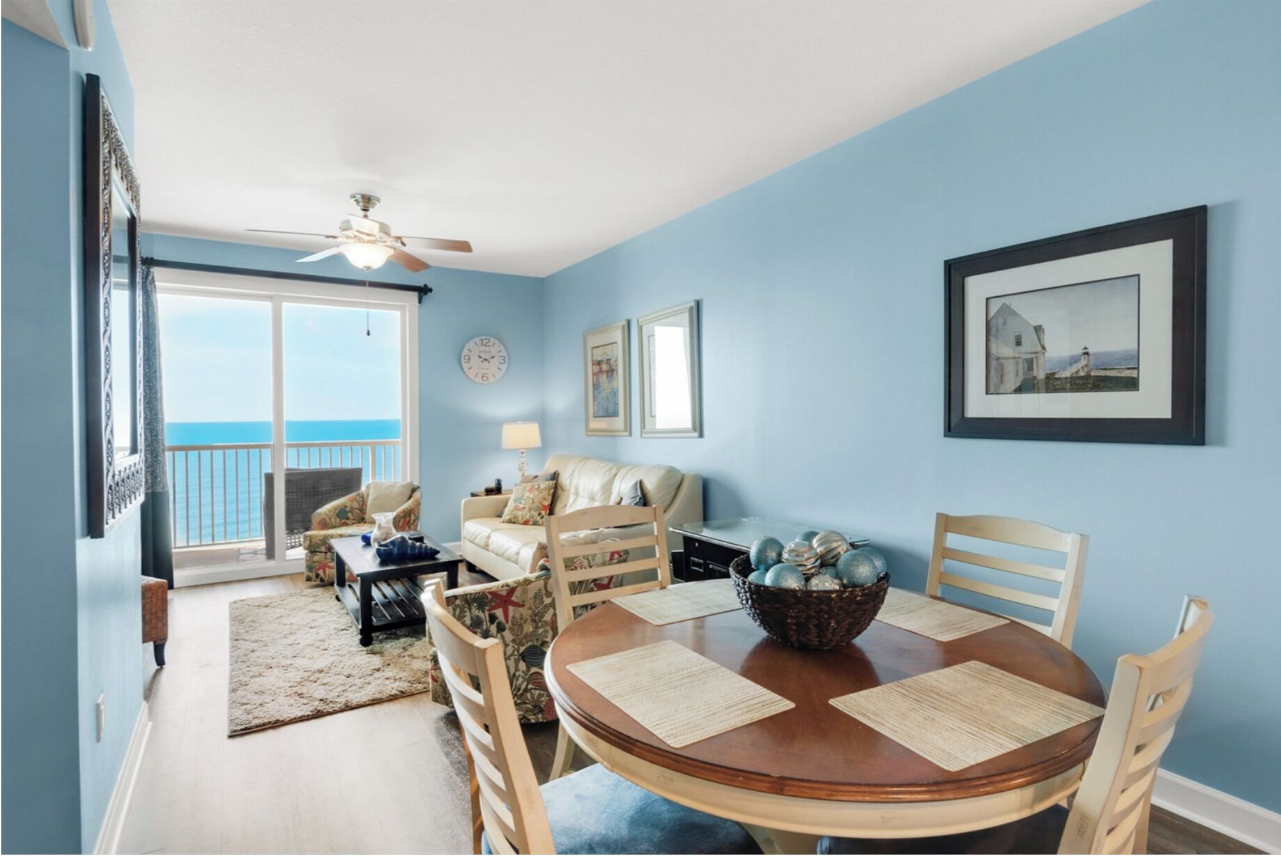A room with a round wooden table, chairs, a sofa, and a balcony with a view.