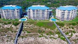 Un complesso alberghiero sulla costa con diversi edifici, ciascuno dotato di balconi e un tetto blu. Un lungo ponte collega gli edifici alla spiaggia.