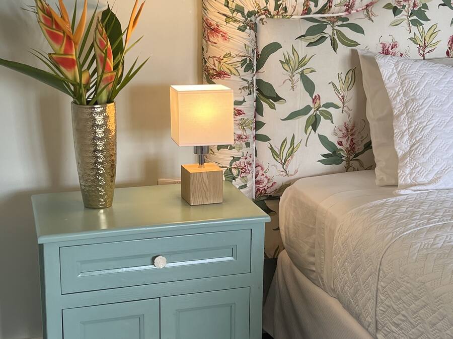 Un dormitorio con cabecera estampada con motivos florales, una mesita de noche con lámpara y una planta decorativa.