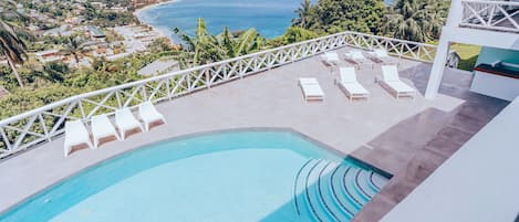 A pool with lounge chairs, a view of the ocean, and a white building with a balcony.