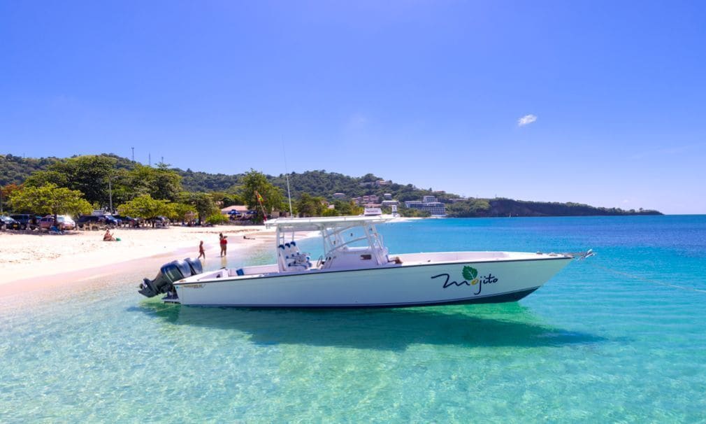 A boat named "Zijitto" docked at a beach with clear turquoise water, a sandy shore, and a backdrop of hills and buildings.