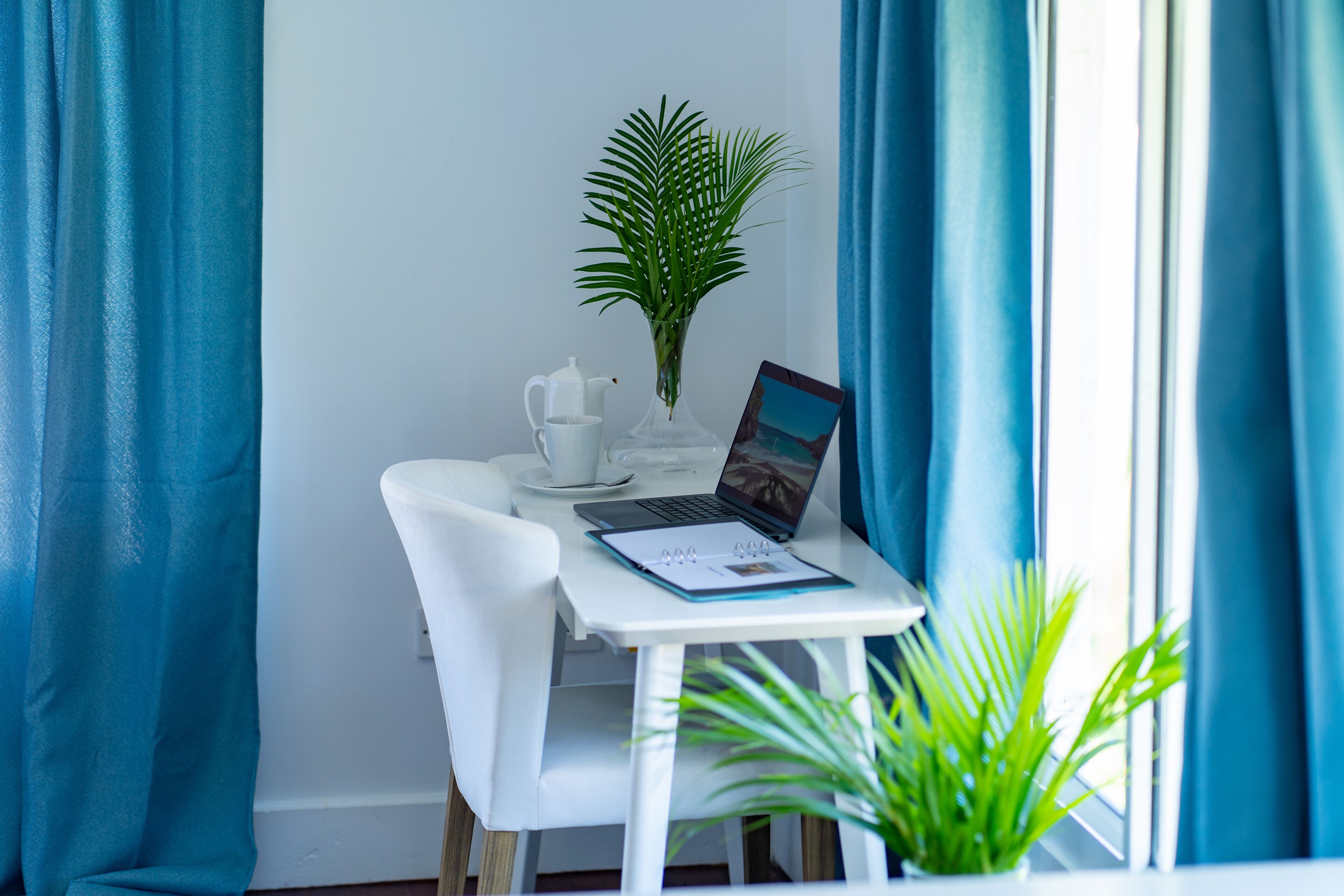 A white table with a laptop, a white mug, a clear vase with a green plant, and a framed picture.