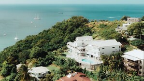 Un edificio grande y blanco con varios balcones y una piscina, rodeado de exuberante vegetación y vistas a un mar tranquilo con varios barcos.