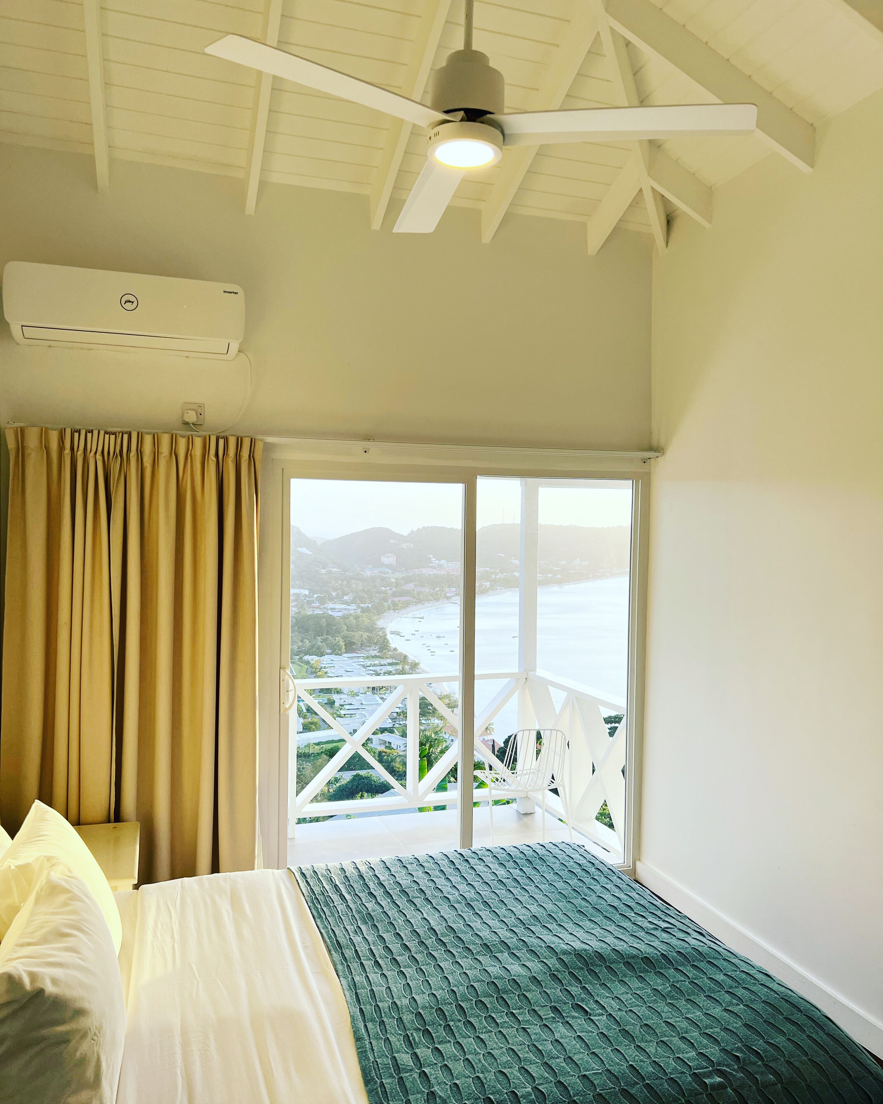 A bedroom with a bed, a ceiling fan, an air conditioning unit, and a large window with a view of a coastal landscape.