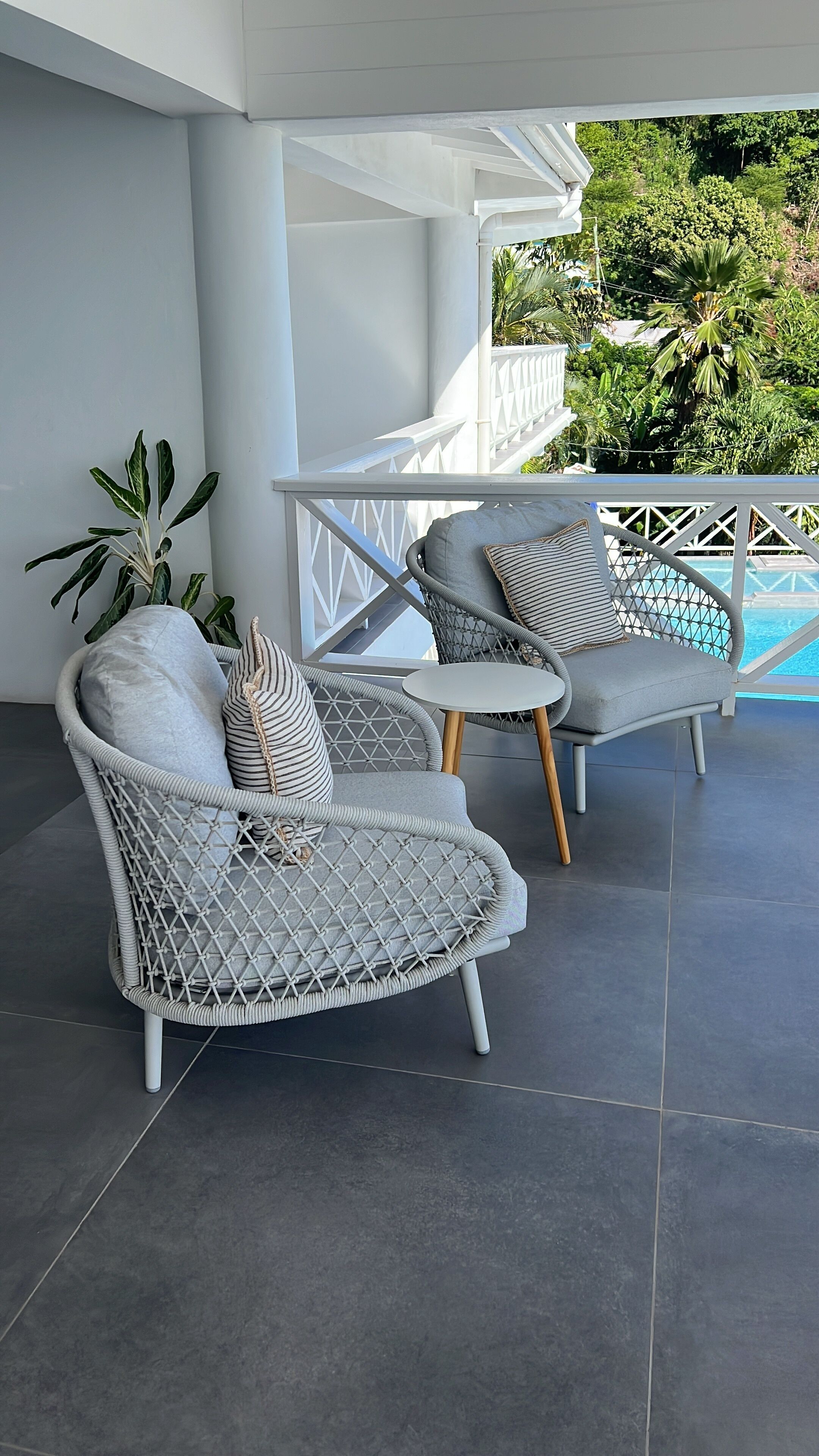 Dos sillones de mimbre con cojines y una pequeña mesa, ubicados en un patio con baldosas y vista a una piscina y follaje.
